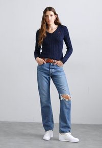 Navy cable-knit sweater with v-neck, paired with light blue distressed jeans. White sneakers complete the outfit. Brown belt adds contrast.