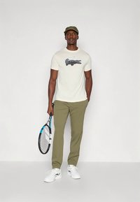 Homem vestindo uma t-shirt branca com um logótipo de crocodilo, calças de tom oliva, sapatilhas brancas e boné, segurando uma raquete de ténis numa mão.