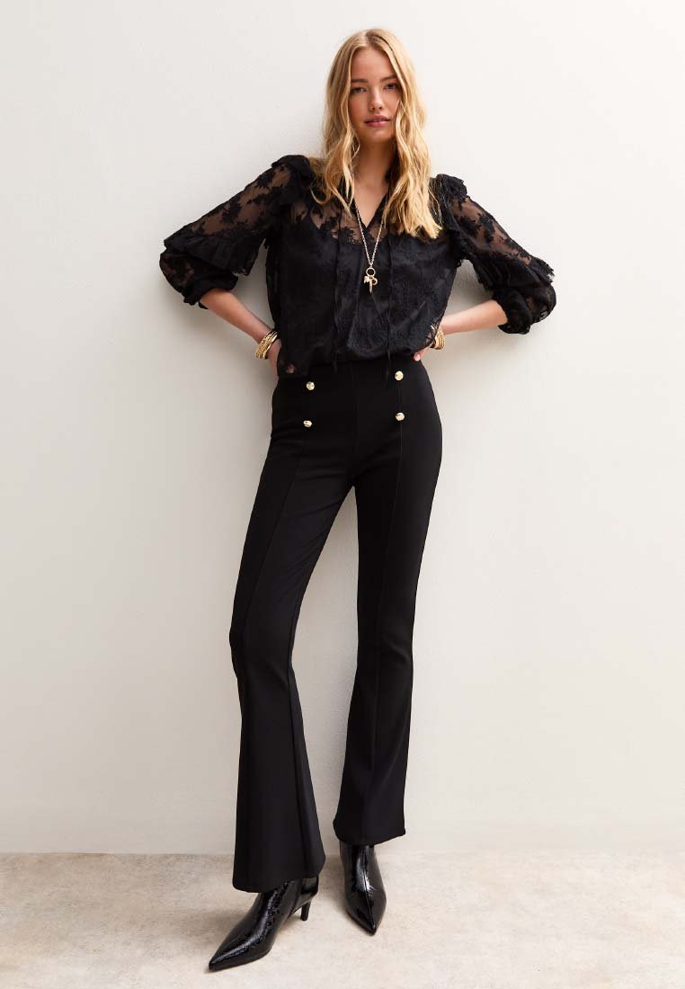 Femme portant un blouse en dentelle florale noire, un pantalon noir taille haute avec des boutons dorés, des bottes noires à bout pointu, se tenant contre un mur blanc.