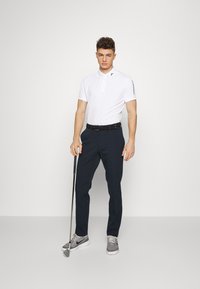 Camisa polo branca com uma lapela de três botões e logotipo no colarinho, combinada com calças de fato azul-marinho e sapatos desportivos cinzentos, segurando um taco de golfe.