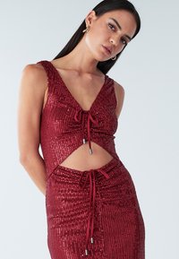 Sarkana samta kleita ar V-veida izgriezumu, izgriezumiem sānos un aukliņu detaļām. Raksturota ar pieguļošu siluetu un teksturētu virsmu.