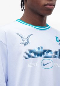 Νέος φορεί λευκό μπλουζάκι με μπλε σήμα Nike, στυλιζαρισμένο πουλί που κρατάει κλαδί ελιάς και γιακά με μπλε ρίγες.