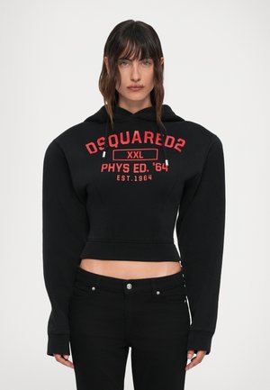 Kvinde iført sort cropped hoodie med rød "DSQUARED2 XXL PHYS ED. '64 EST.1964" tekst og sorte bukser, stående mod hvid baggrund.