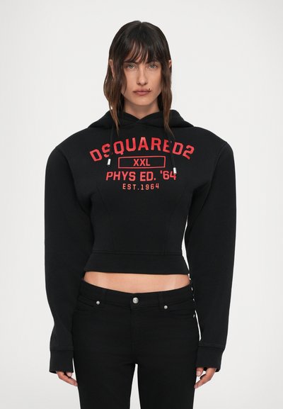 Femeie îmbrăcată cu un hanorac negru scurt cu textul roșu "DSQUARED2 XXL PHYS ED. '64 EST.1964" și pantaloni negri, stând pe un fundal alb.
