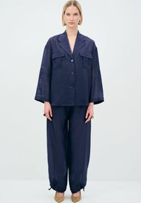Giacca oversize di lino blu navy con due tasche frontali, abbinata a pantaloni larghi coordinati. Dettagli dei bottoni notevoli e tessuto testurizzato.
