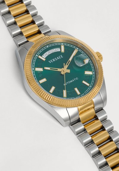 Montre automatique de luxe Versace avec cadran vert, bracelet métal or et argent, lunette dorée, affichage du jour et de la date, et index des heures luminescents.