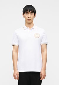 Άνδρας που φοράει λευκό πόλο με κοντά μανίκια, γιακά και μανίκια με χρυσή διακόσμηση, και στρογγυλό λογότυπο Versace Jeans Couture στο αριστερό στήθος.