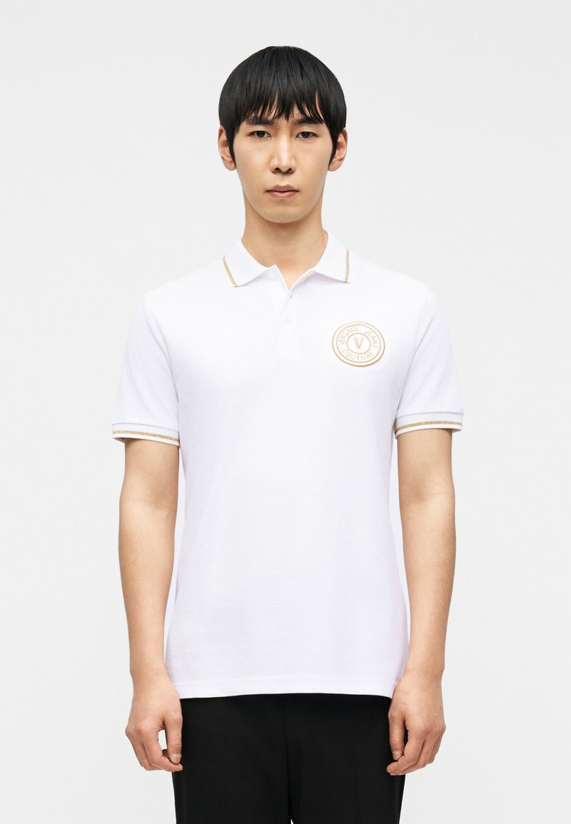 Άνδρας που φοράει λευκό πόλο με κοντά μανίκια, γιακά και μανίκια με χρυσή διακόσμηση, και στρογγυλό λογότυπο Versace Jeans Couture στο αριστερό στήθος.