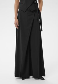 Vrouw in zwarte wijde wrap-broek met een riem, gecombineerd met zwarte puntige schoenen, op een witte achtergrond.