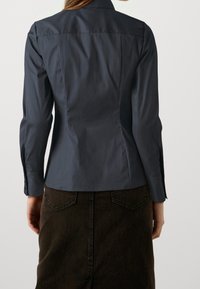 Donna che indossa una maglietta a maniche lunghe grigio scuro attillata e una gonna di jeans nera a vita alta, vista di spalle su uno sfondo semplice.