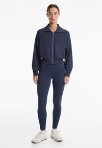 Navy Zip-Front-Jacke mit breitem Kragen und strukturiertem Stoff, kombiniert mit navy Leggings und weißen Sneakers. Beide Kleidungsstücke haben eine taillierte Passform.