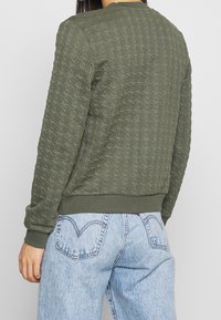 Olivgrön vadderad sweatshirt med texturerat mönster, ribbade ärmslut och nederkant, tillsammans med ljusblå högmidjade jeans.