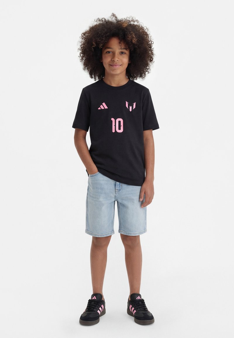 Αγόρι με σγουρά μαλλιά, φοράει μαύρο μπλουζάκι με ροζ λογότυπα και τον αριθμό 10, ανοιχτόχρωμο τζιν σορτς και μαύρα αθλητικά παπούτσια με ροζ ρίγες.