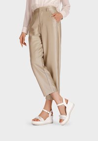 Pantaloni beige in satin a vita alta con tasche laterali, abbinati a sandali bianchi con zeppa, dotati di cinturino alla caviglia e fibbia decorativa.