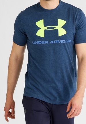Osoba w niebieskiej koszulce Under Armour z dużym żółtym logo i ciemnych sportowych spodniach na jednolitym jasnym tle.