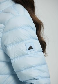 Giacca puffer di colore blu chiaro con texture trapuntata, dotata di un patch con logo di montagna nero sulla manica sinistra. Materiale morbido e liscio.