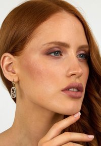 Orecchino a cerchio in oro con un ciondolo rettangolare decorativo caratterizzato da un design intricato. La modella ha lunghi capelli rossi e un trucco neutro.