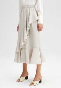 Falda beige claro con detalles de volantes, cintura alta y dobladillo acampanado, hecha de tela suave y texturizada. Combinada con sandalias de tacón de color claro.