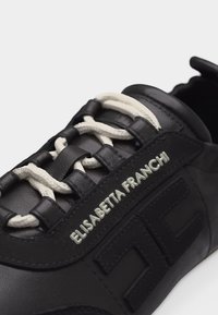 Μαύρο δερμάτινο αθλητικό παπούτσι με λευκά κορδόνια και κεντημένο το "ELISABETTA FRANCHI" στο πλάι κοντά στα ανάγλυφα αρχικά της μάρκας.