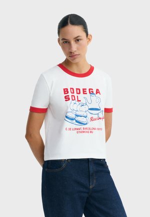 Kobieta ubrana w białą koszulkę z czerwonym wykończeniem, zdobioną niebieskimi i czerwonymi ilustracjami jedzenia oraz napojów i napisem, w połączeniu z ciemnoniebieskimi dżinsami.