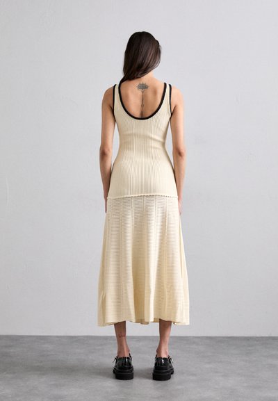 Vestido de punto acanalado en color crema con un cuerpo ajustado y una falda acampanada en capas. Presenta una espalda baja y un ribete negro en contraste en los hombros.