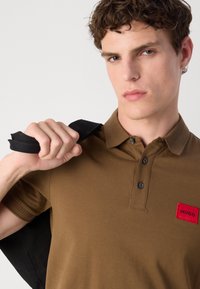 Tricou polo maro cu două butoane, guler ribbit și un patch roșu cu logo-ul "HUGO". Modelul ține o geantă de umăr neagră cu o mână.