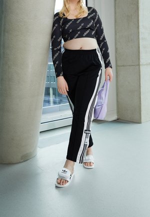 Black long-sleeve crop top with repeated "adidas" logo, black pants with white stripes, and white slide sandals. Purple bag held in one hand.