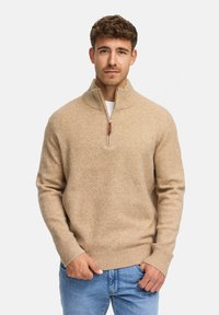 Beiger Strickpullover mit hohem Kragen und halbseitigem Reißverschluss. Weiche Textur und gerippter Saum, kombiniert mit hellblauen Jeans.
