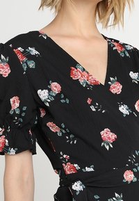 Svart klänning med v-ringning och puffärmar, med ett blommönster i röd, rosa och vit. Texturerad tyg med en knytning i midjan.