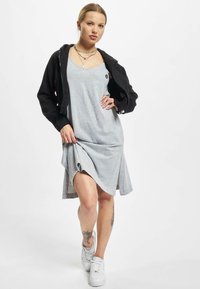 Grå ermeløs kjole med asymmetrisk kant, lagd under en svart gå-opp-hettegenser. Modellen har på seg hvite joggesko og viser frem legentatoveringer.