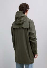 Person som bär en olivgrön huvjacka, sedd bakifrån, stående mot en enfärgad ljusgrå bakgrund.