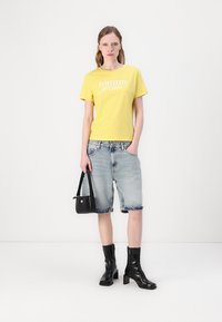 Gul bomull t-skjorte med "Tommy Jeans"-logo, kombinert med denim Bermuda-shorts og svarte hælboots. Svart veske med logo-detalj.