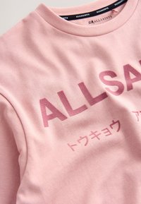 Vaaleanpunainen collegepaita, valmistettu pehmeästä puuvillasta, jossa on suuri tummanpunainen logoteksti ja japanilaiset merkit sen alla.