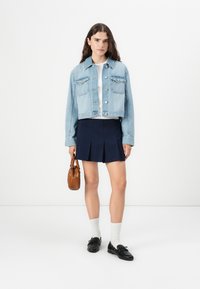 Lys blå denim cropped jakke med to brystlommer, kombinert med et marineblått plissert skjørt, hvite sokker, svarte loafers og en brun håndveske.