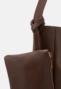 Sac à main en cuir marron foncé avec détail de lanière nouée et petite pochette zippée attachée, présenté en gros plan sur fond neutre.