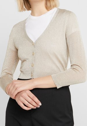 Femme portant un cardigan court beige scintillant avec des boutons sur une chemise blanche, associé à un pantalon noir taille haute.