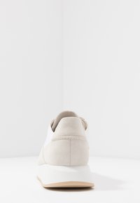 Sneaker grigio chiaro con accenti bianchi, materiale scamosciato, forma arrotondata e suola bianca spessa. La vista posteriore mostra un design minimal e dettagli di cucitura.