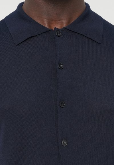 Primo piano di una persona di pelle scura che indossa una polo in maglia blu navy con colletto a piatto e bottoni.