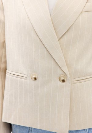 Blazer beige à fines rayures, croisé, à revers crantés, deux boutons et poches passepoilées, porté sur un jean bleu clair.