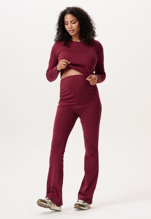 Vrouw met krullend haar die een bijpassende bordeauxrode top met lange mouwen en een broek met hoge taille draagt, waarbij ze de zoom van haar shirt iets optilt.