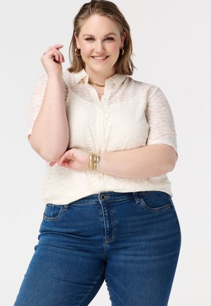 Lachende vrouw met een witte kanten korte mouwen blouse en een blauwe spijkerbroek, poseert met armen over elkaar en gouden sieraden tegen een effen achtergrond.
