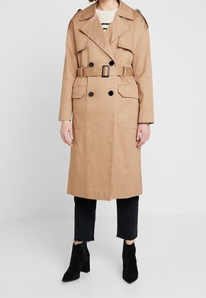 Kobieta w beżowym trenczu z paskiem, na białym swetrze, czarne spodnie do kostek i czarne botki, stojąca na jednolitym tle.