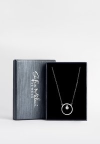 Sølv halskæde præsenteret i en sort fløjlsæske. Indeholder en cirkulær vedhæng med en lille sten i midten. Æsken har en struktureret grå finish.