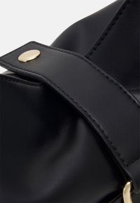 Svart läderväska med slät textur, försedd med en bred rem som hålls på plats av en guldfärgad knappar. Sömmen är synlig.