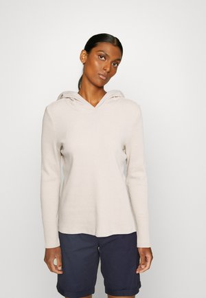 Helle beige Kapuzenjacke mit langen Ärmeln, gerippter Textur und V-Ausschnitt. Kombiniert mit dunkelblauen Shorts für einen Kontrast.