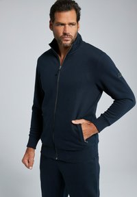 Chaqueta de forro polar azul marino con cuello alto, detalles de bolsillos y puños de canalé. Fabricada en una tela suave y con un diseño ajustado, es adecuada para el uso casual.