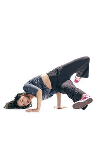 Sportbekleidung: ein geknotetes graues Oberteil und locker sitzende schwarze Hose, kombiniert mit rosa Turnschuhen. Die Figur befindet sich in einer Breakdance-Pose.