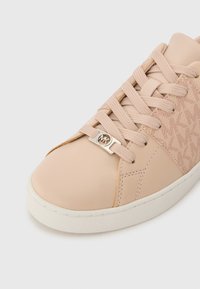 Beige sneaker met een gestructureerd, gepatterned zijpaneel; glad bovenwerk; platte veters; metalen logo-accent; rubberen zool; minimalistisch ontwerp.