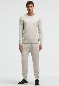 Helles beiges Langarmshirt mit Rundhalsausschnitt und strukturiertem Stoff, kombiniert mit passenden beigen Jogginghosen und schwarzen Turnschuhen.