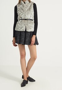 Grå faux pälsväst med hög krage, bälte i midjan, bärs över en svart långärmad topp och en svängig svart kjol med ett texturerat mönster.